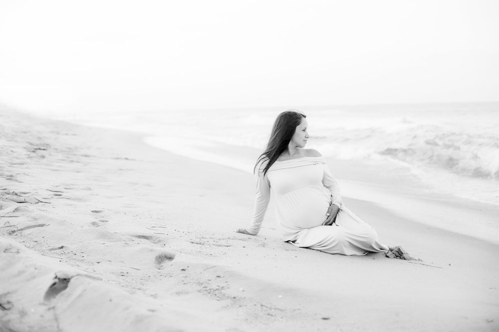 Black and white image of pregnant mom sitting on the beach looking out at the ocean.