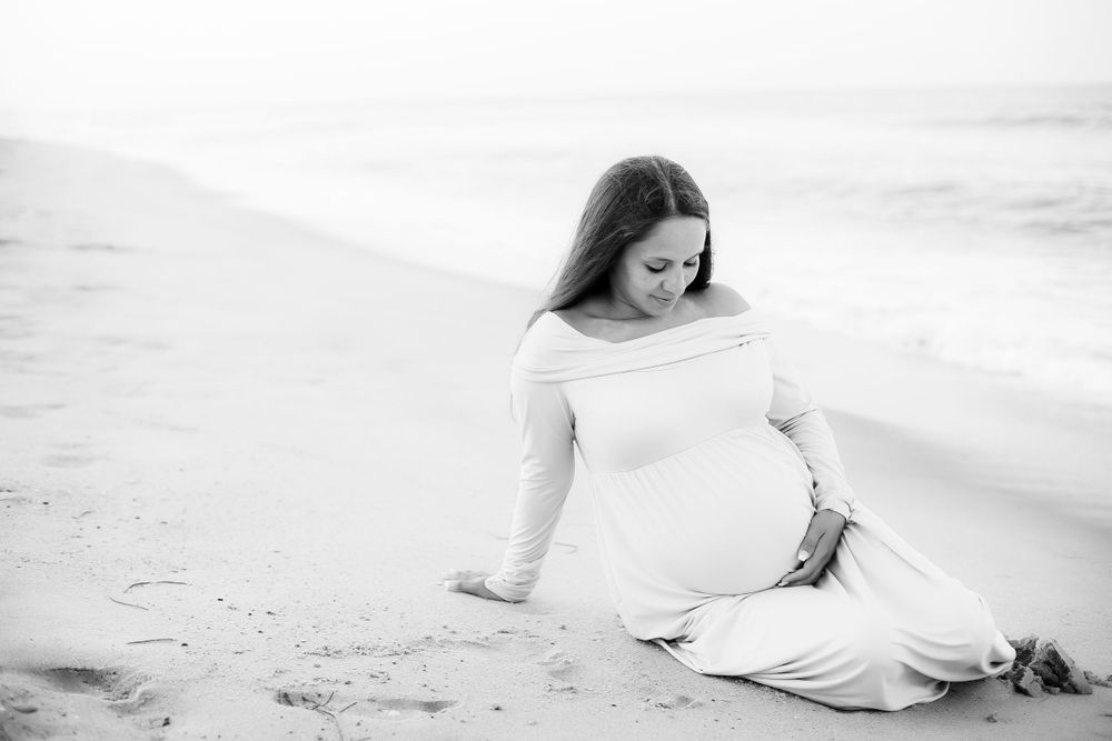 Black and white image of pregnant mom sitting on the beach gazing down at her belly.