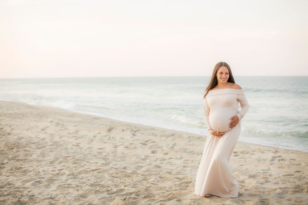 Pregnant mom in a white dress standing on the beach holding her belly with a beautiful sunset behind her.