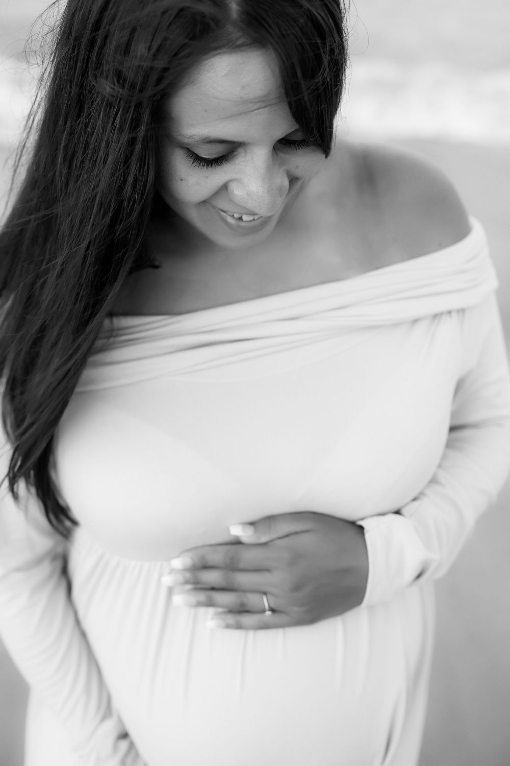 Black and white image of pregnant woman standing on the beach in white dress with hand showing her ring.