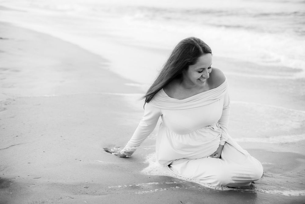 Black and white image of pregnant woman sitting in the surf on the beach.  She is laughing as the water hit her legs.