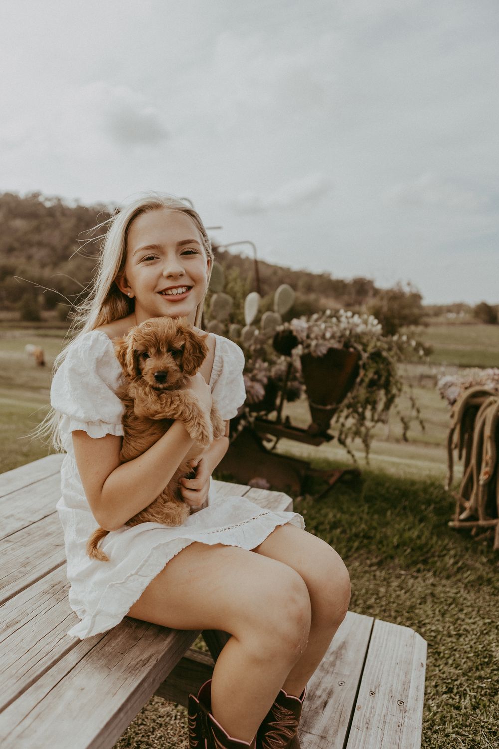 A smiling blonde girl in a white dress sits on a wooden bench holding an adorable fluffy red puppy.