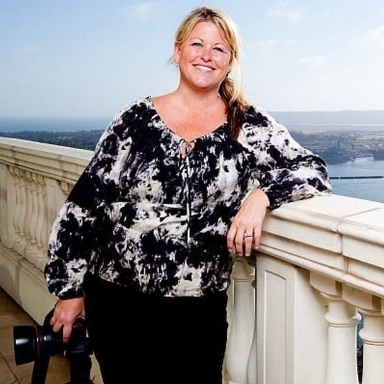 Woman in black and white tie-dye blouse posing on balcony overlooking water.