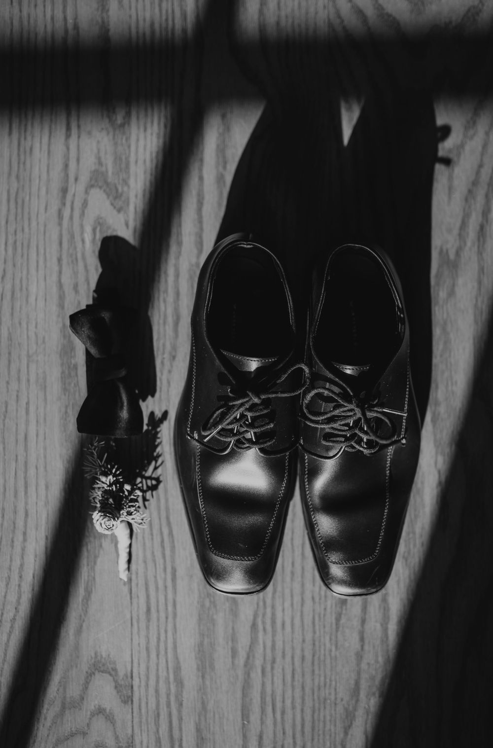 Black and white flat lay of groom's dress shoes, bow tie, and boutonniere on wooden floor.
