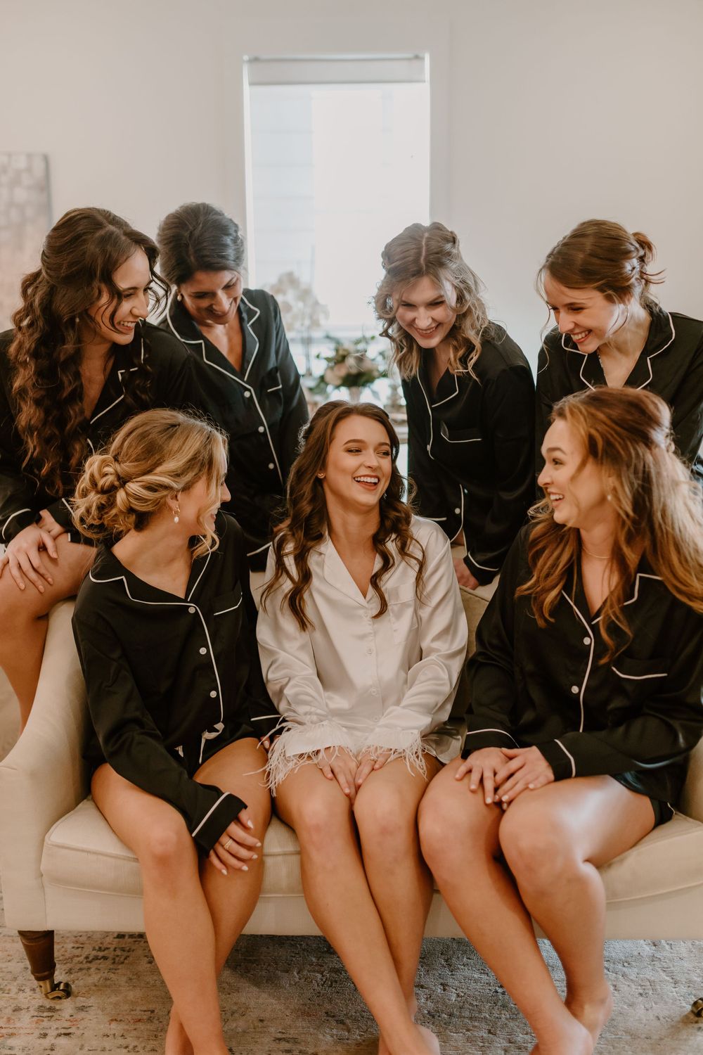 Bride in white robe surrounded by bridesmaids in black pajamas laughing during wedding morning prep.
