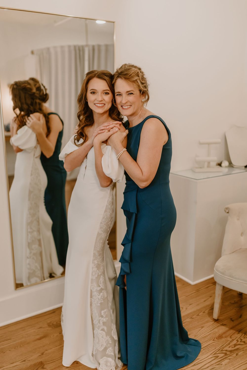 Bride in lace wedding gown posing with woman in teal dress in front of mirror.