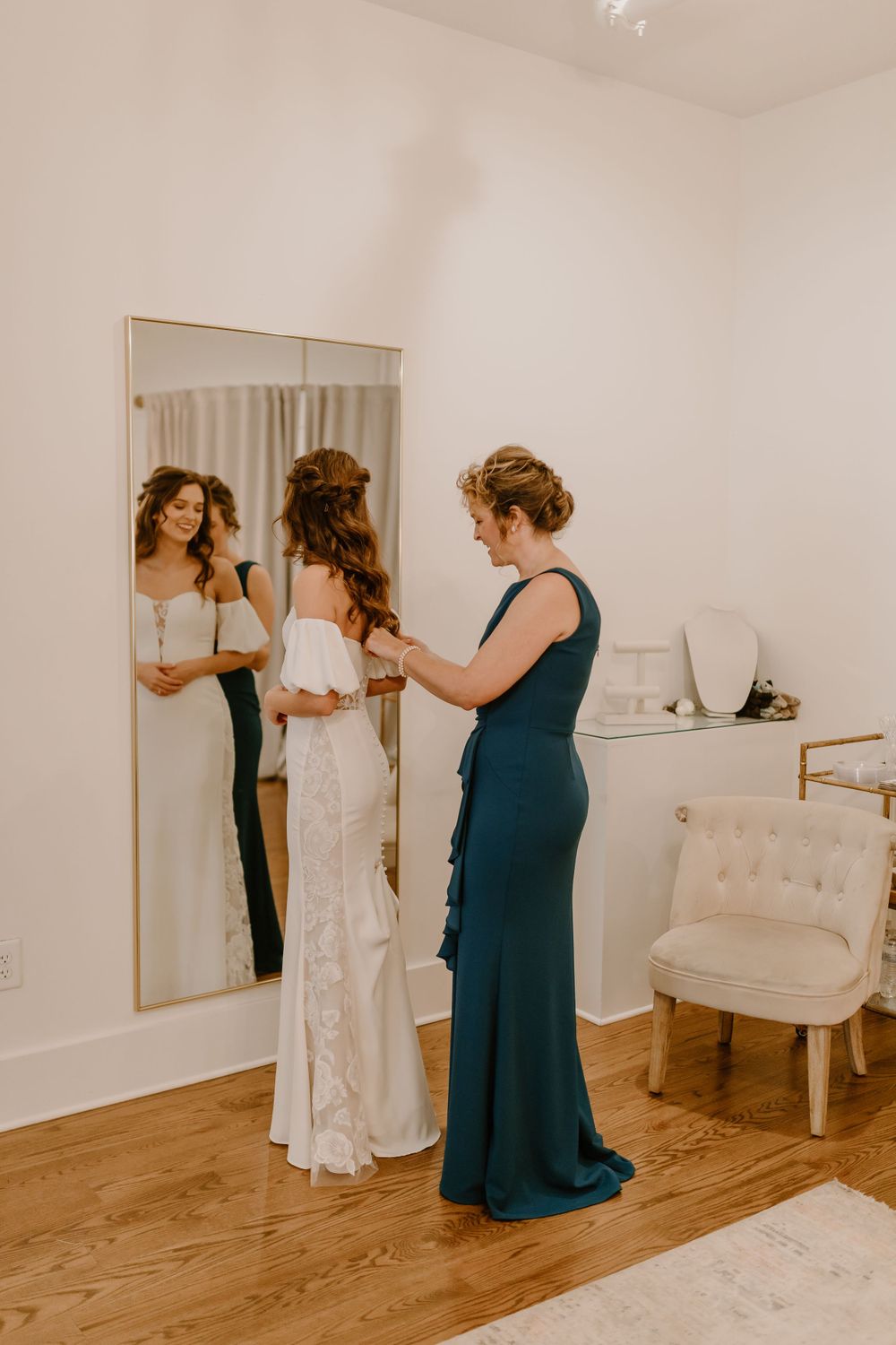 Mother helping bride button her off-shoulder lace wedding gown in bright white dressing room with mirror reflection.