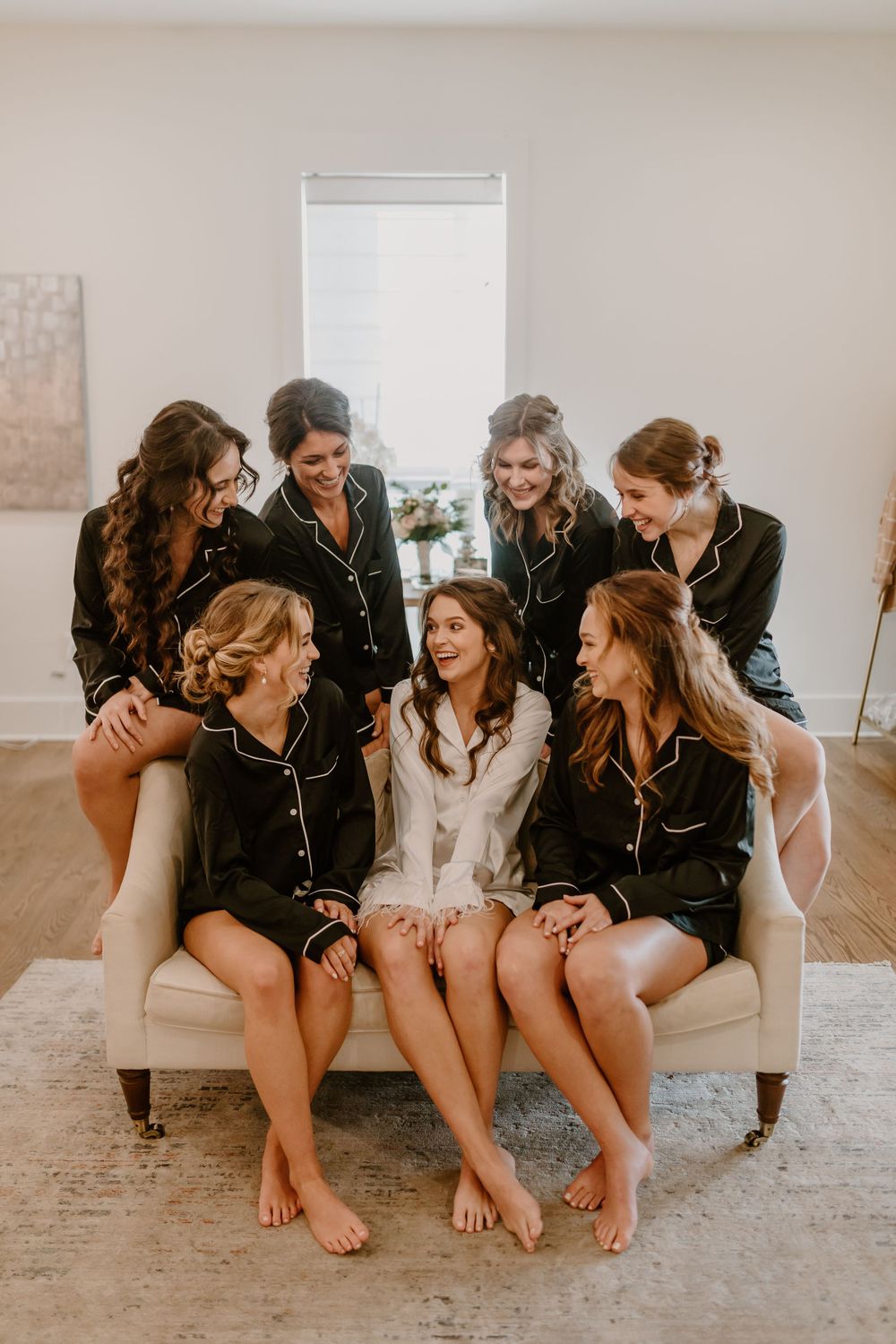 Bride surrounded by six bridesmaids in matching black pajamas sitting together on cream sofa, laughing and smiling.