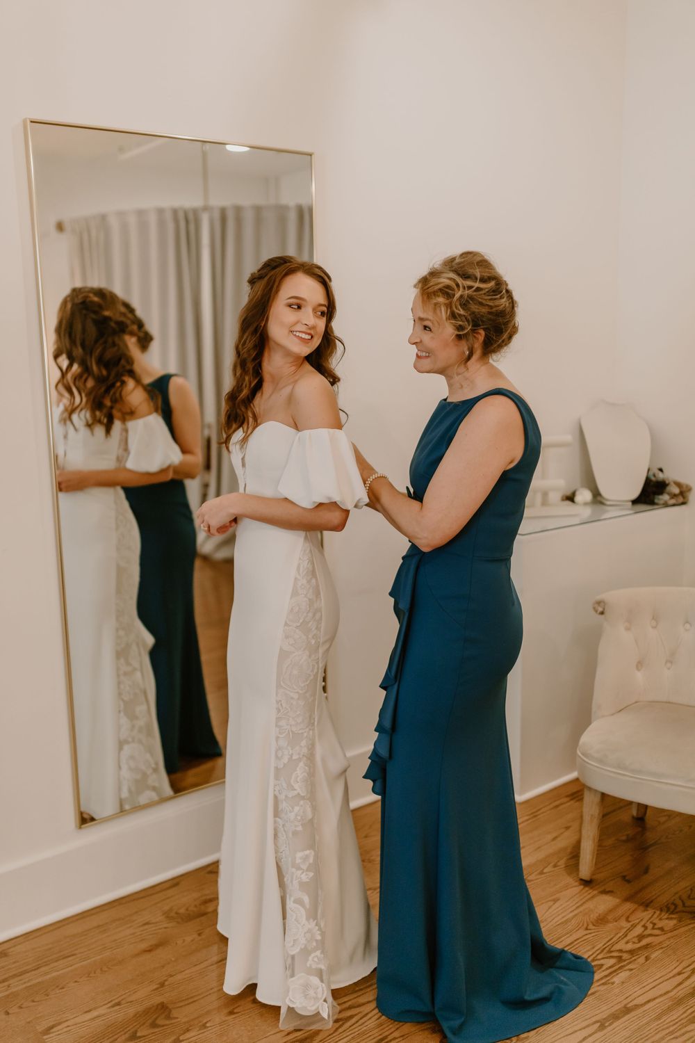 Smiling bride in elegant off-shoulder wedding dress sharing a moment with her mother in a bright dressing room.