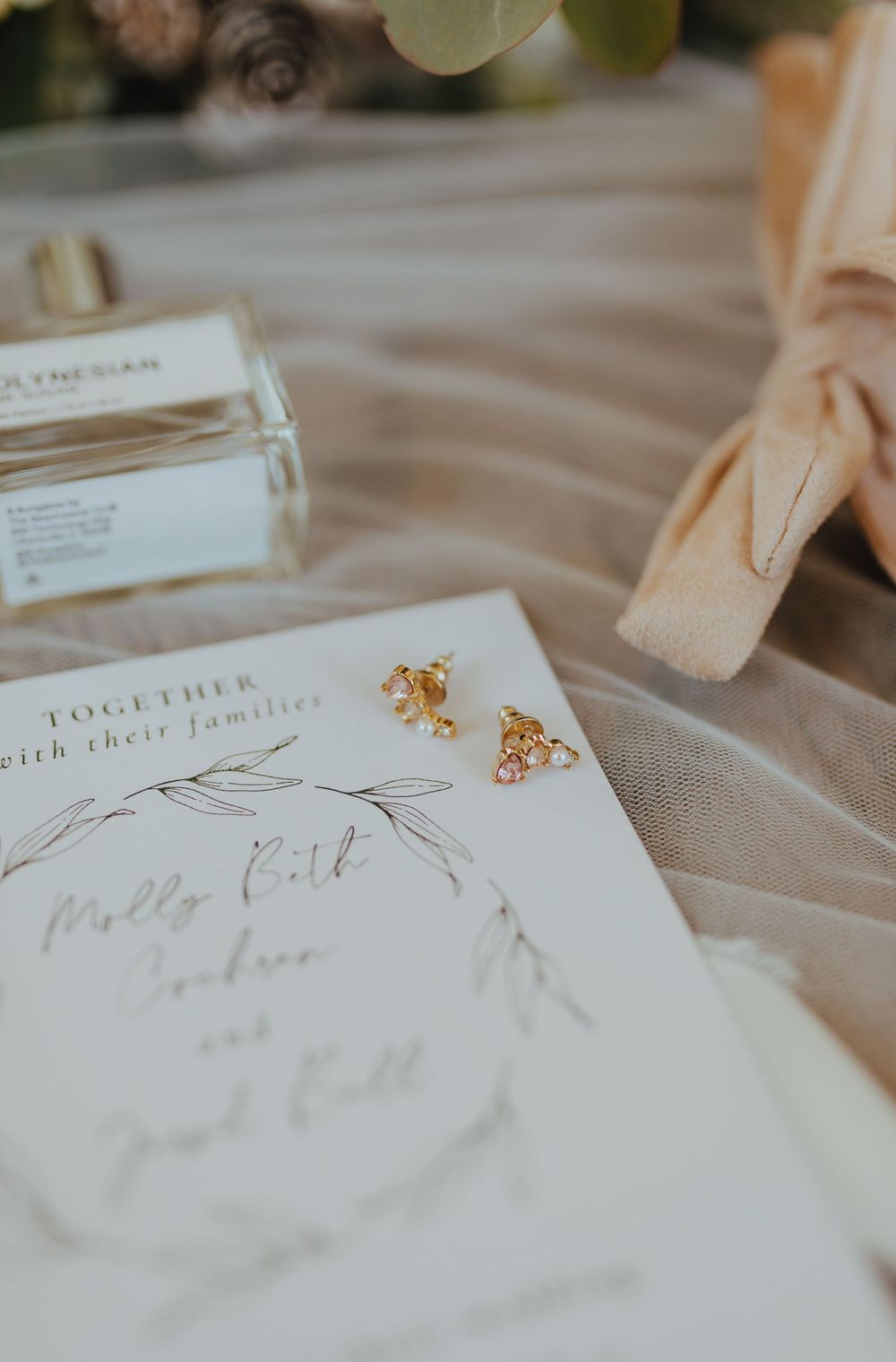Wedding invitation with calligraphy, gold floral earrings, perfume bottle, and blush ribbon on tulle.