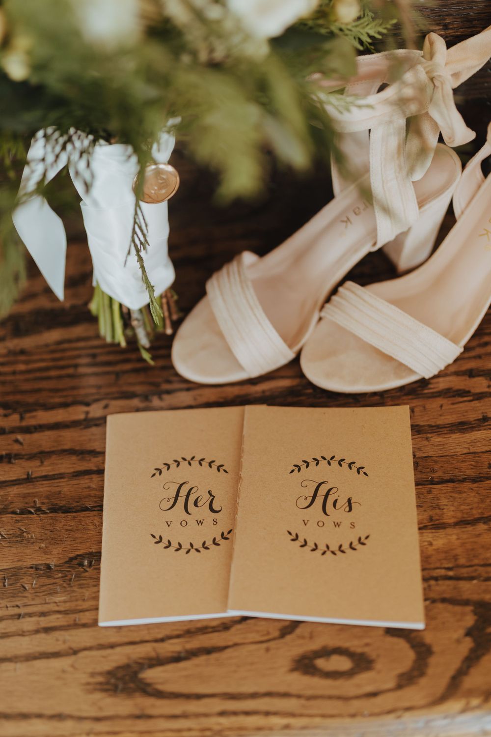 His and Her kraft paper vow booklets beside white bridal heels and bouquet on rustic wooden surface.