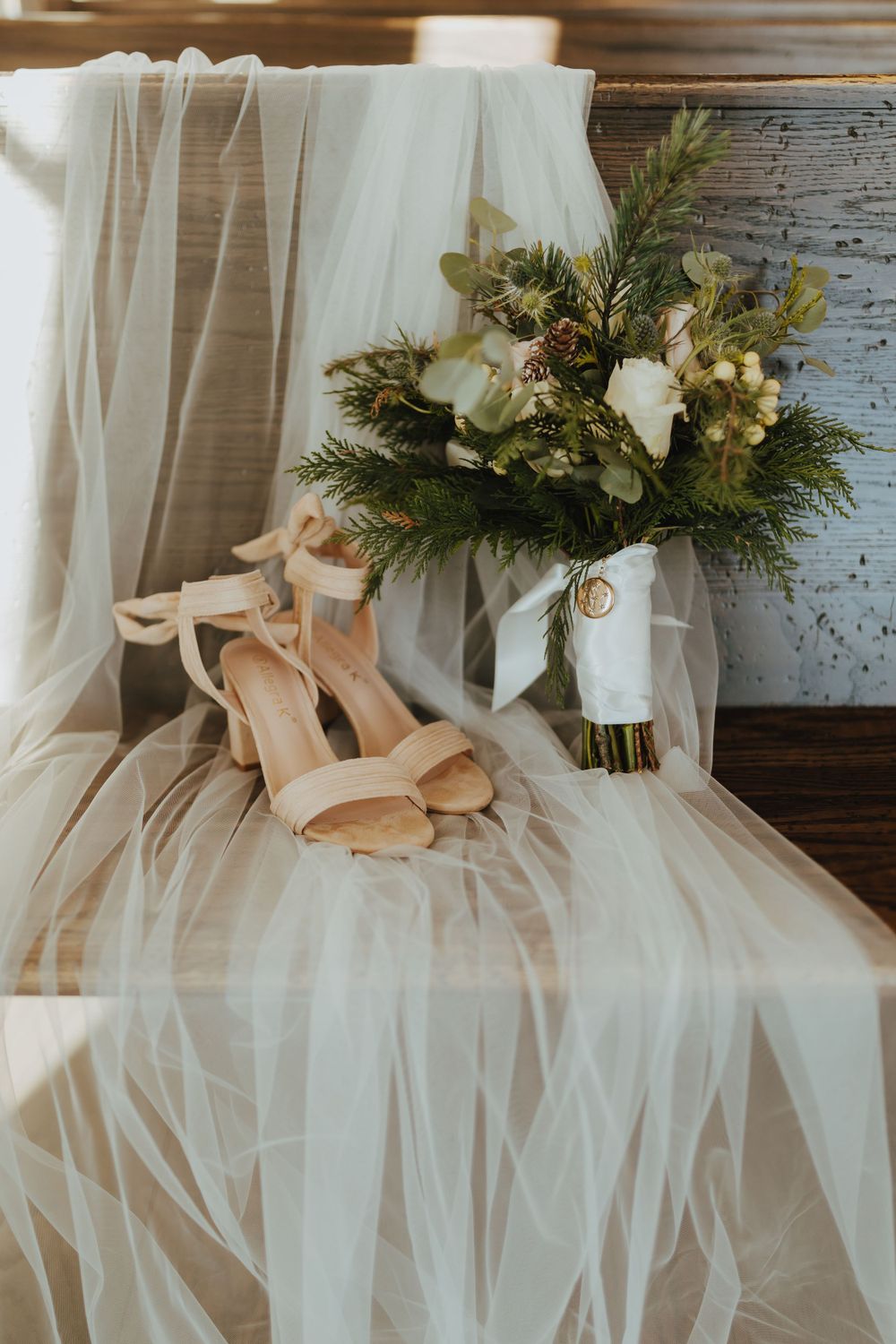 Nude ankle-strap heels and winter bridal bouquet with white roses and pine on draped white tulle.