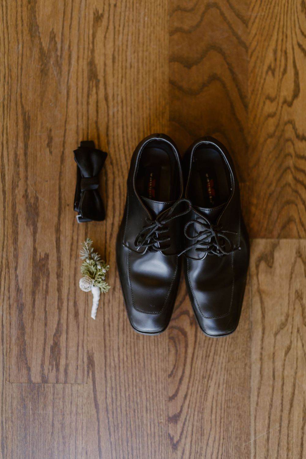 Black leather Oxford dress shoes with bow tie and floral boutonniere on wooden floor for groom.