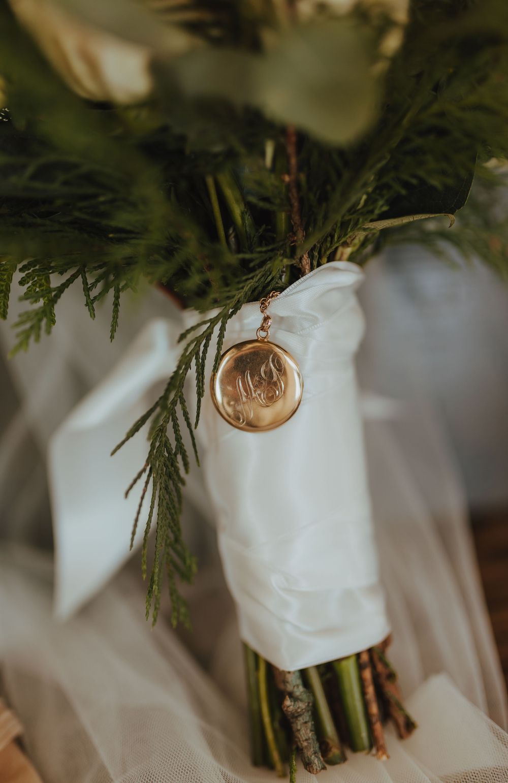 Bridal bouquet with evergreen foliage wrapped in white ribbon with gold monogram locket charm.