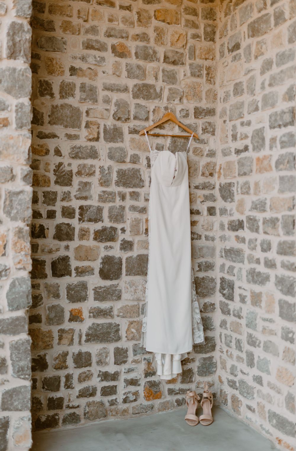 Elegant white wedding dress hanging on wooden hanger against rustic stone wall with nude heels below.