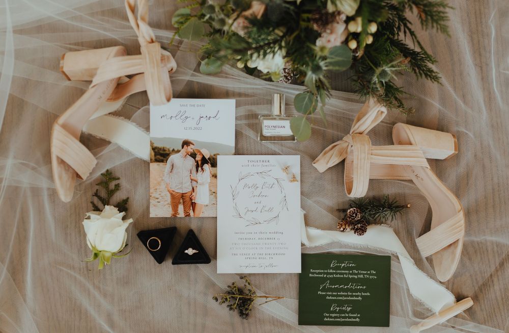 Flat lay of wedding stationery, heels, rings, boutonniere, and greenery arranged on white tulle fabric.