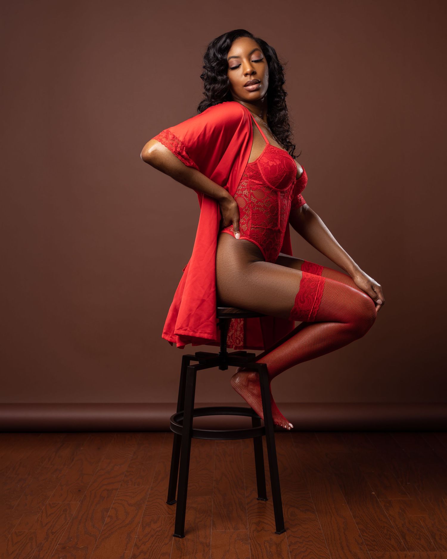Model in red lace bodysuit poses elegantly on wooden stool against brown background.