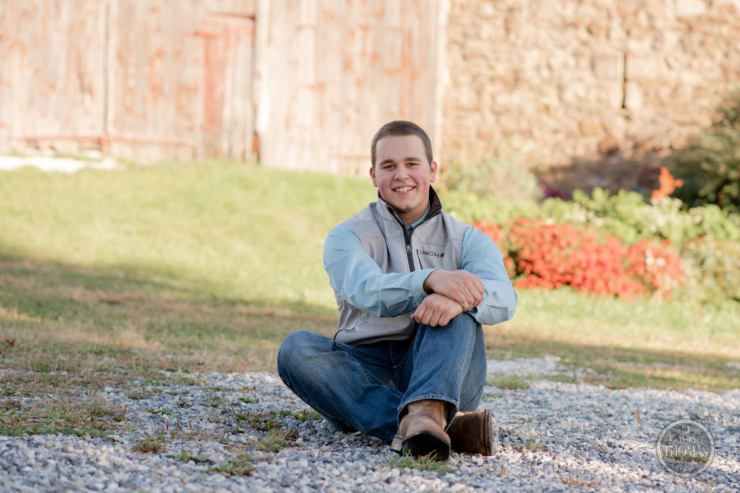 Person in casual attire sits cross-legged on gravel with barn in background.