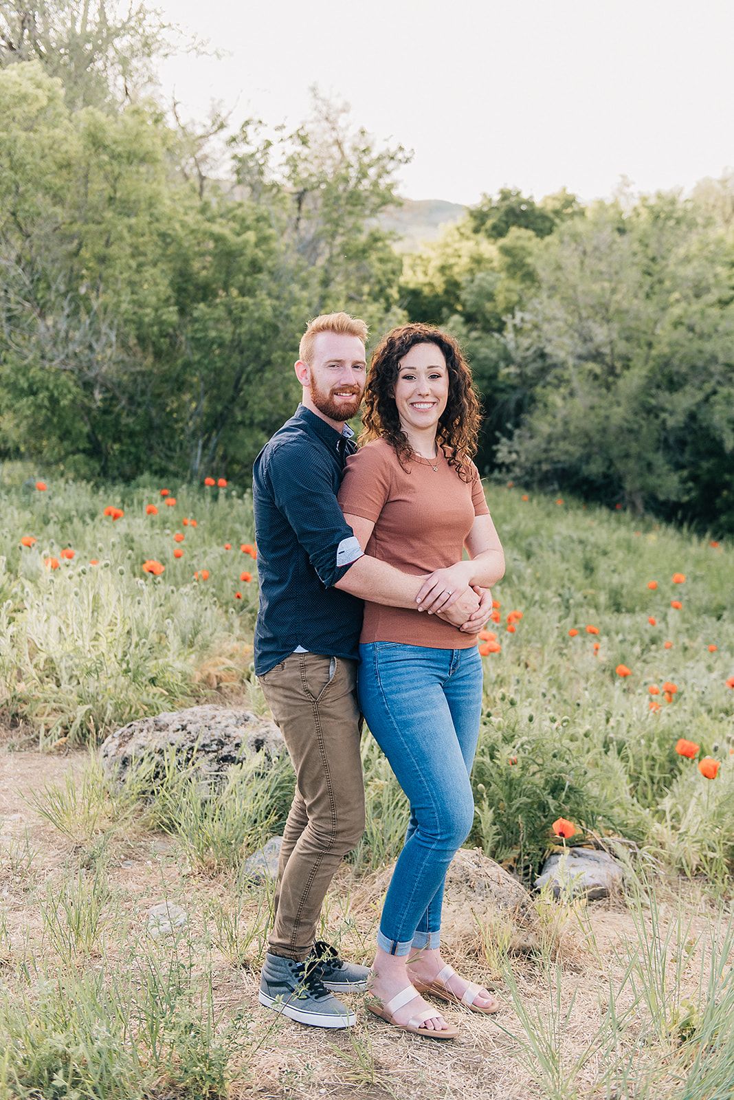 A loving embrace captured in a wildflower meadow with orange poppies and green trees.
