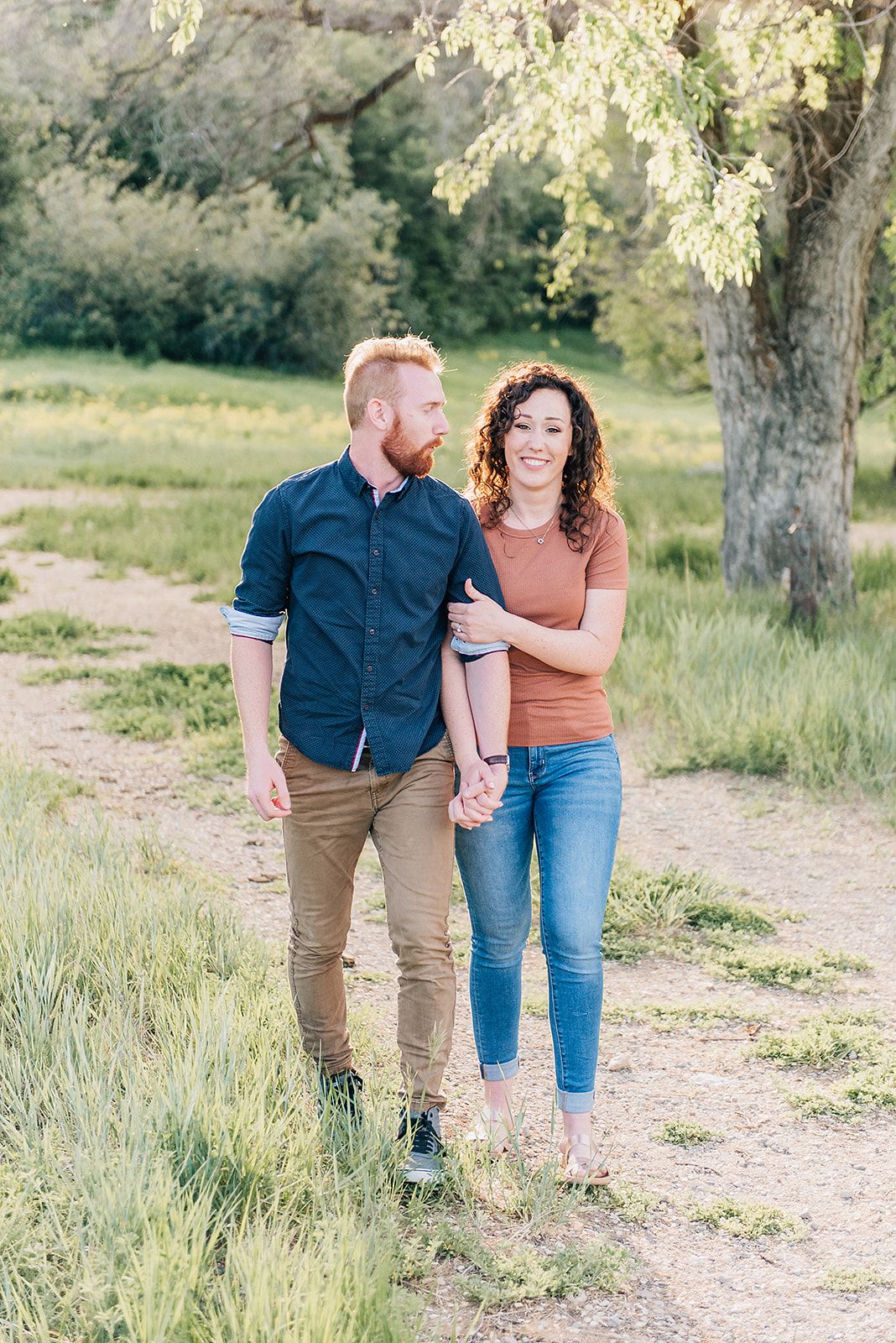 A couple walks together on a nature trail surrounded by green grass and trees.