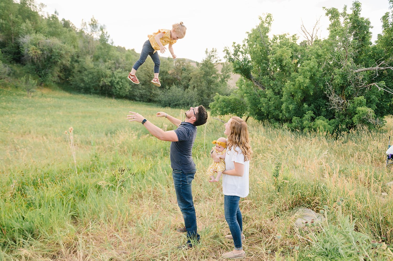 A joyful outdoor moment with a family tossing a baby in the air against a natural landscape background.