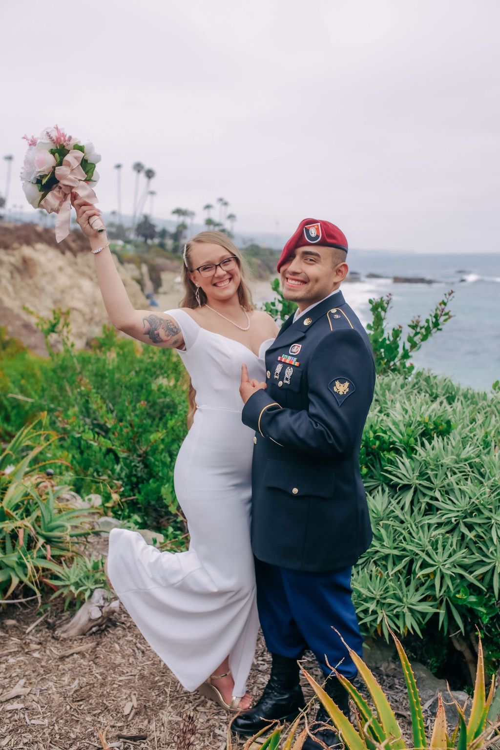 Military wedding couple celebrating on coastal cliff with bouquet and formal attire.
