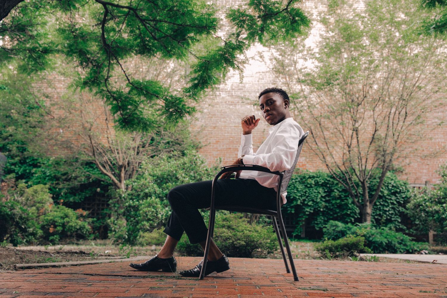 A young Black man in a white shirt sits thoughtfully on a chair in a lush outdoor garden setting.