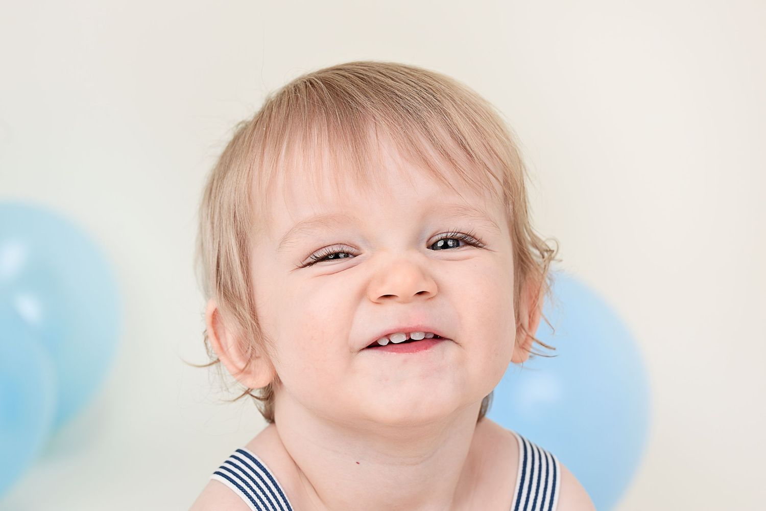 Toddler in striped outfit smiling with blue balloons in background during portrait session.