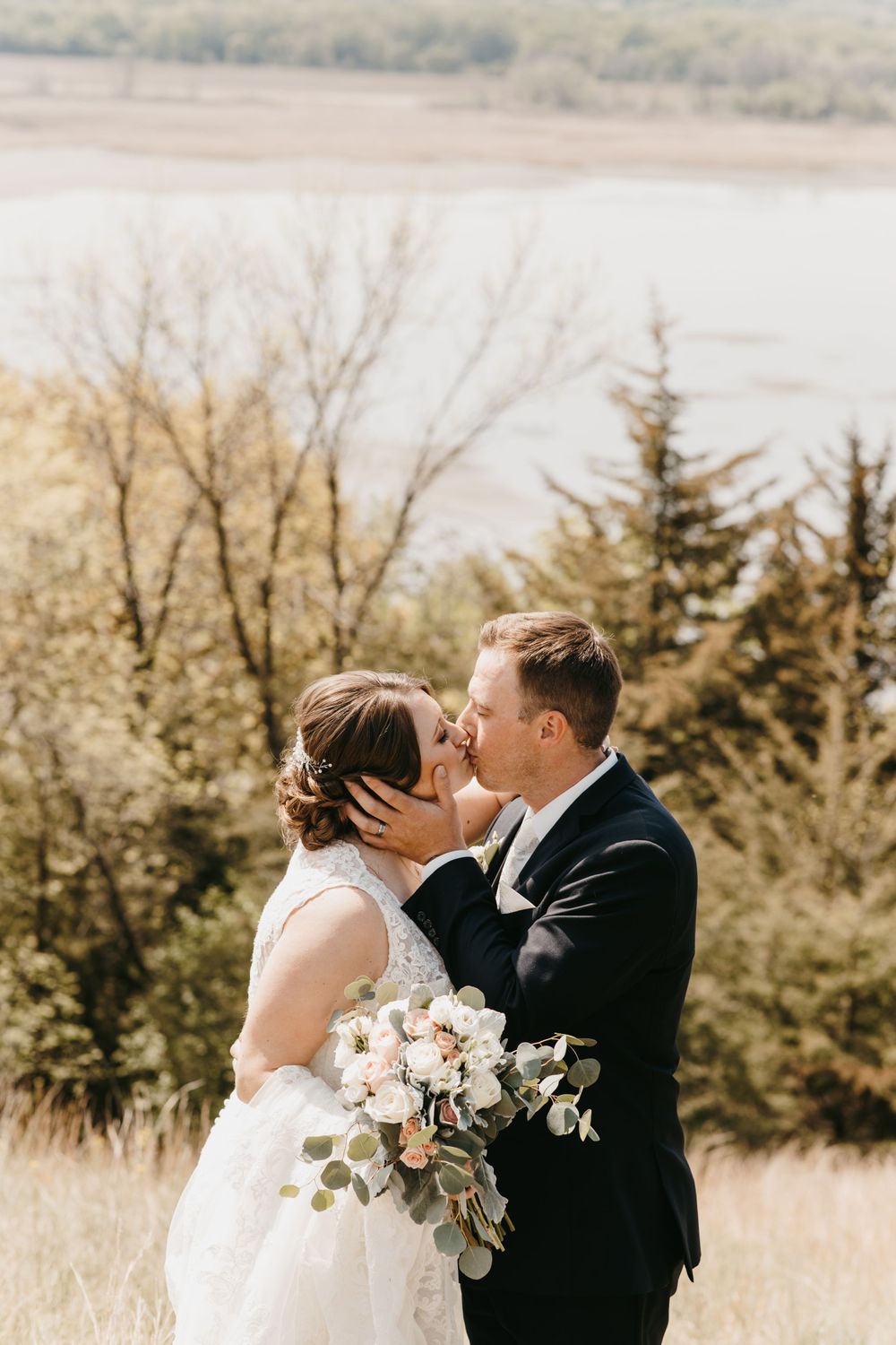 Bride and groom sharing a romantic kiss outdoors with a river and trees in the background on their wedding day.
