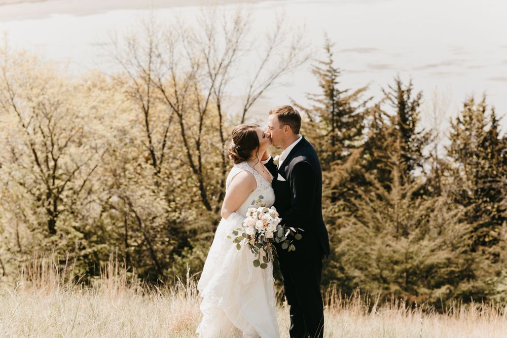 Wedding couple standing close together in a grassy field with evergreen trees and a lake visible behind them.