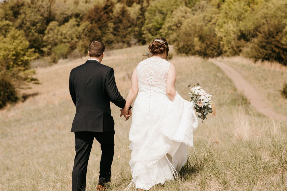 Newlyweds holding hands walking away through a golden grass field, bride carrying a white bouquet.