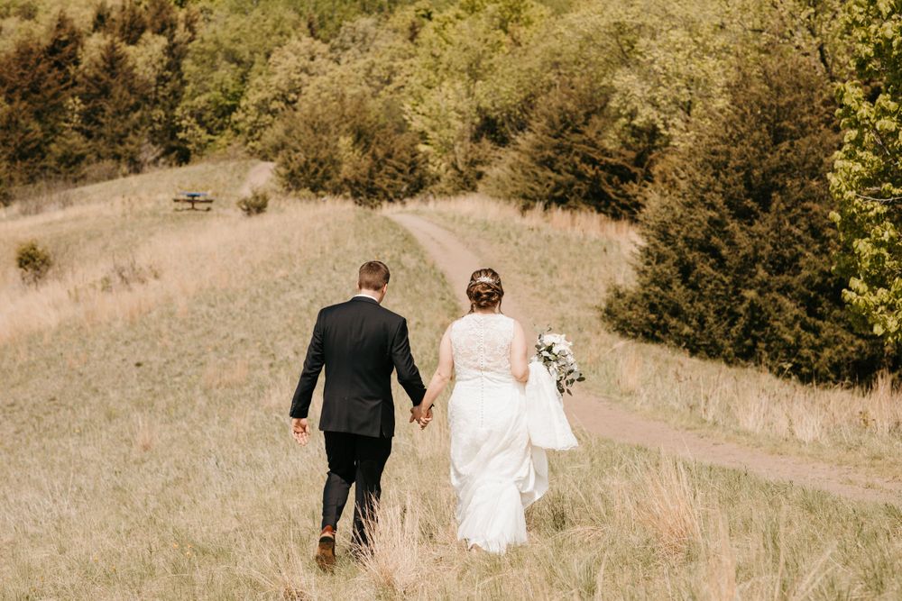 Bride and groom walking hand in hand through a sunny meadow path surrounded by lush green trees.