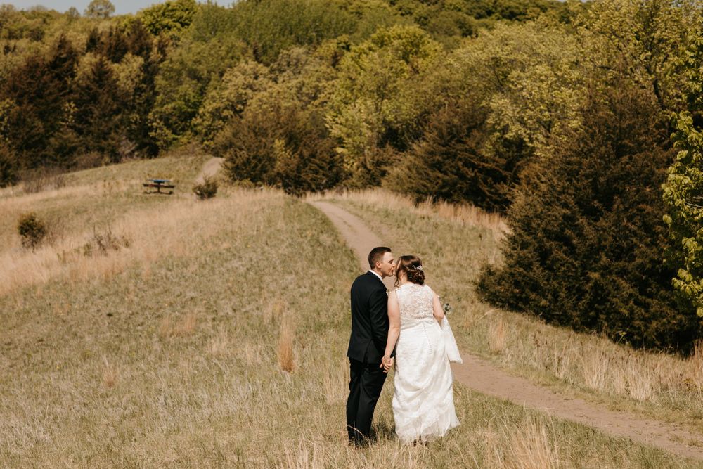 Bride and groom sharing a kiss on a scenic meadow path with dense green trees in the background.