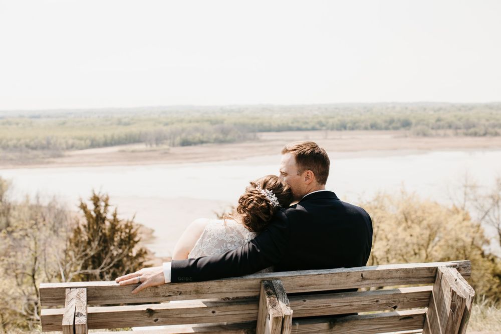 Bride and groom sitting closely together on a rustic wooden bench overlooking a river landscape.