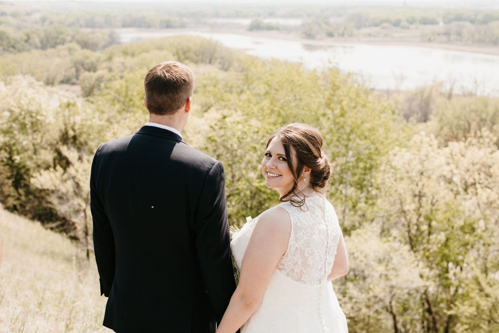 Smiling bride glances back at camera while standing with groom overlooking a scenic river valley.