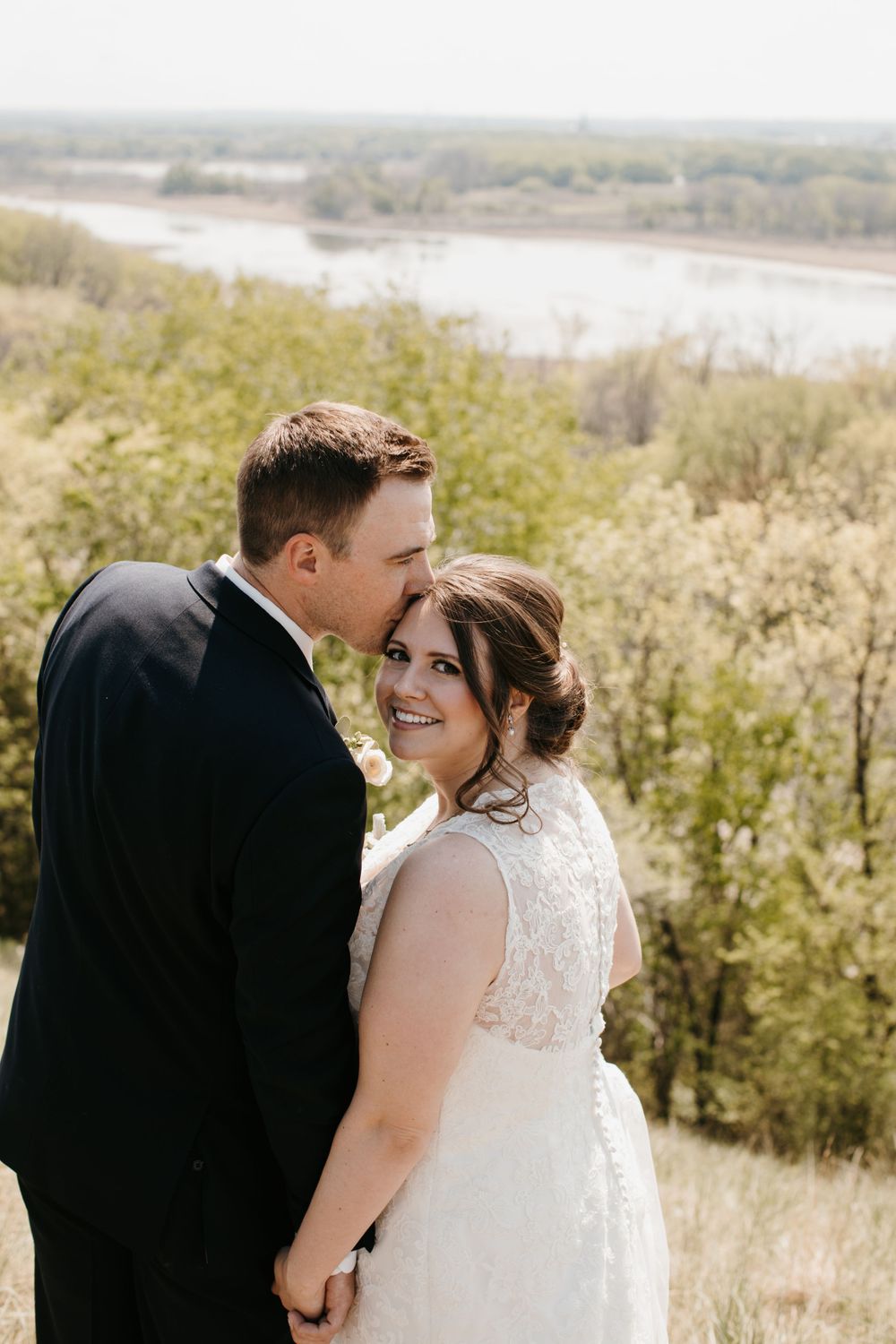 Groom tenderly kissing bride on the cheek as she smiles, with a river valley backdrop behind them.