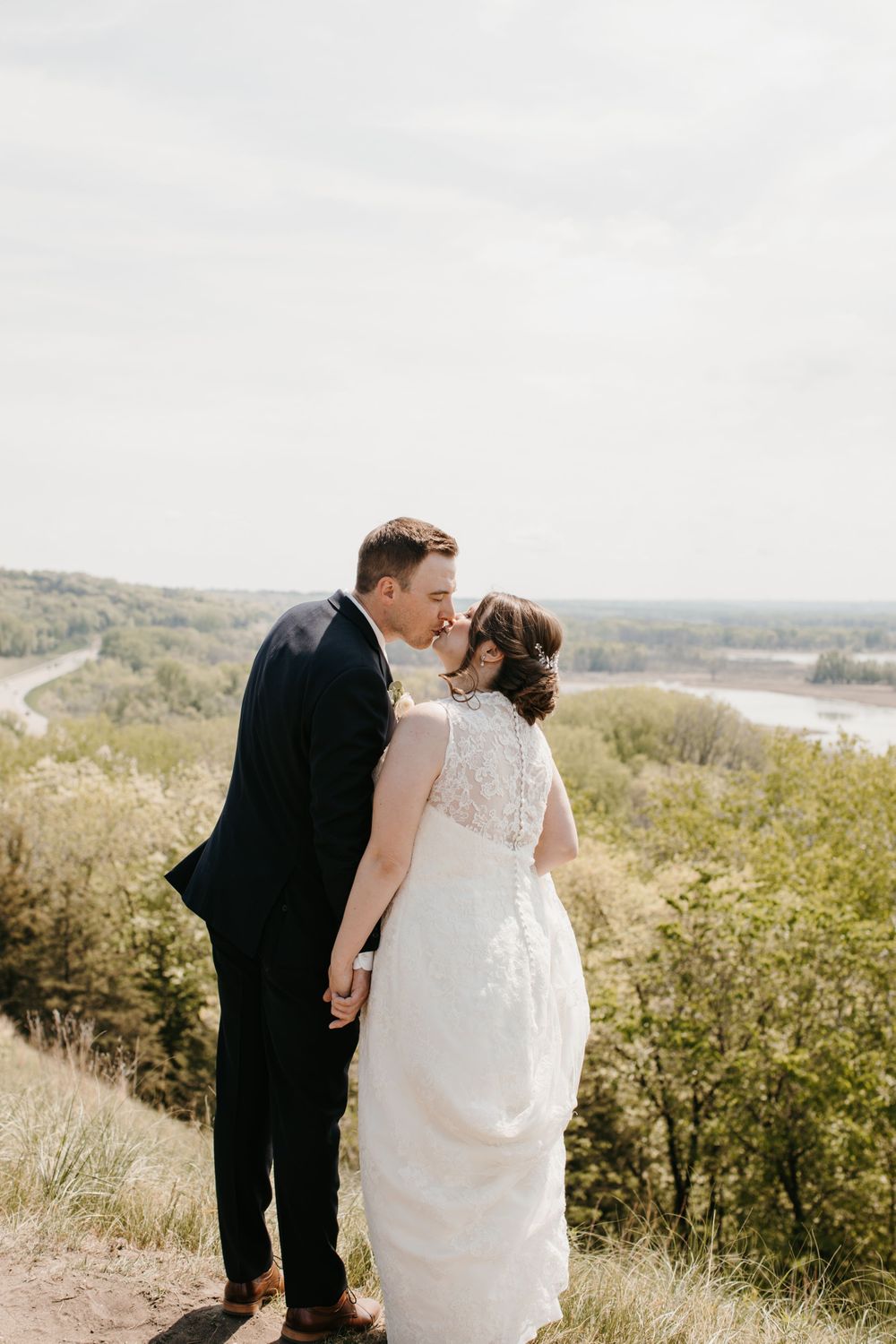Bride and groom kissing on a hilltop with a winding river and lush green landscape stretching below.