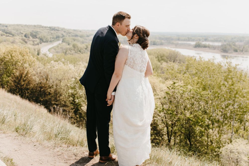 Newlywed couple sharing a kiss on a scenic overlook with a river and rolling hills in the background.