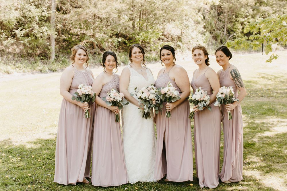Bride in white lace gown poses with five bridesmaids in dusty rose gowns holding floral bouquets outdoors.