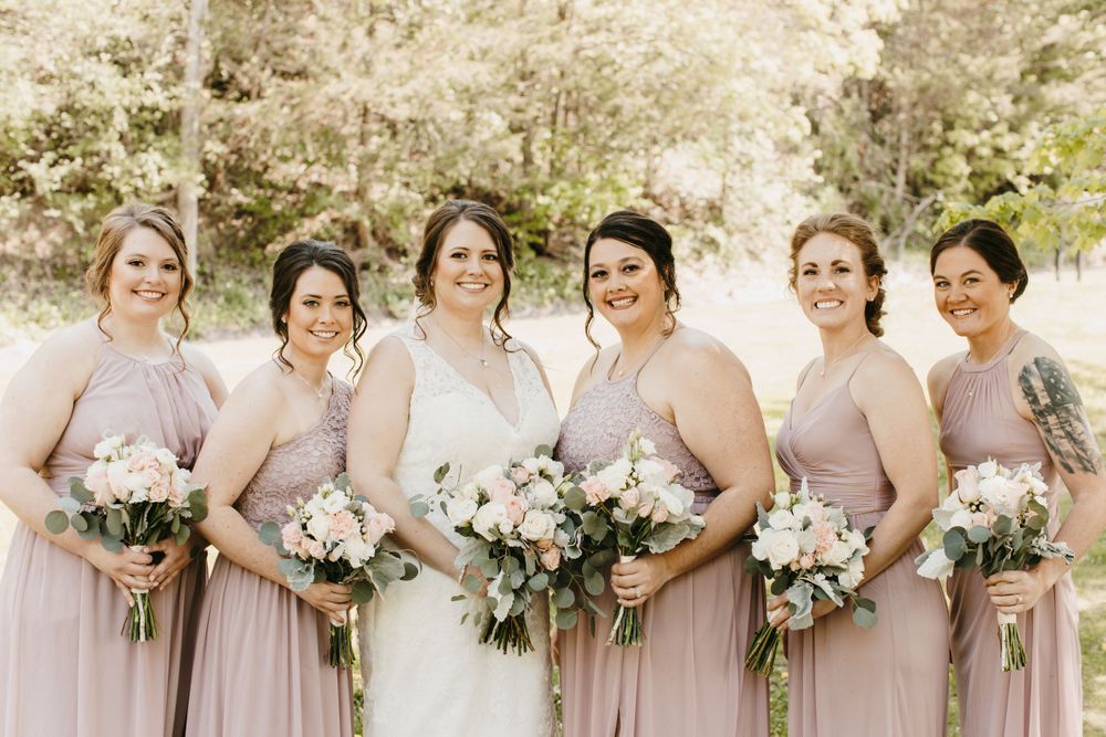 Smiling bride and five bridesmaids in mauve dresses hold white and blush bouquets in a garden setting.