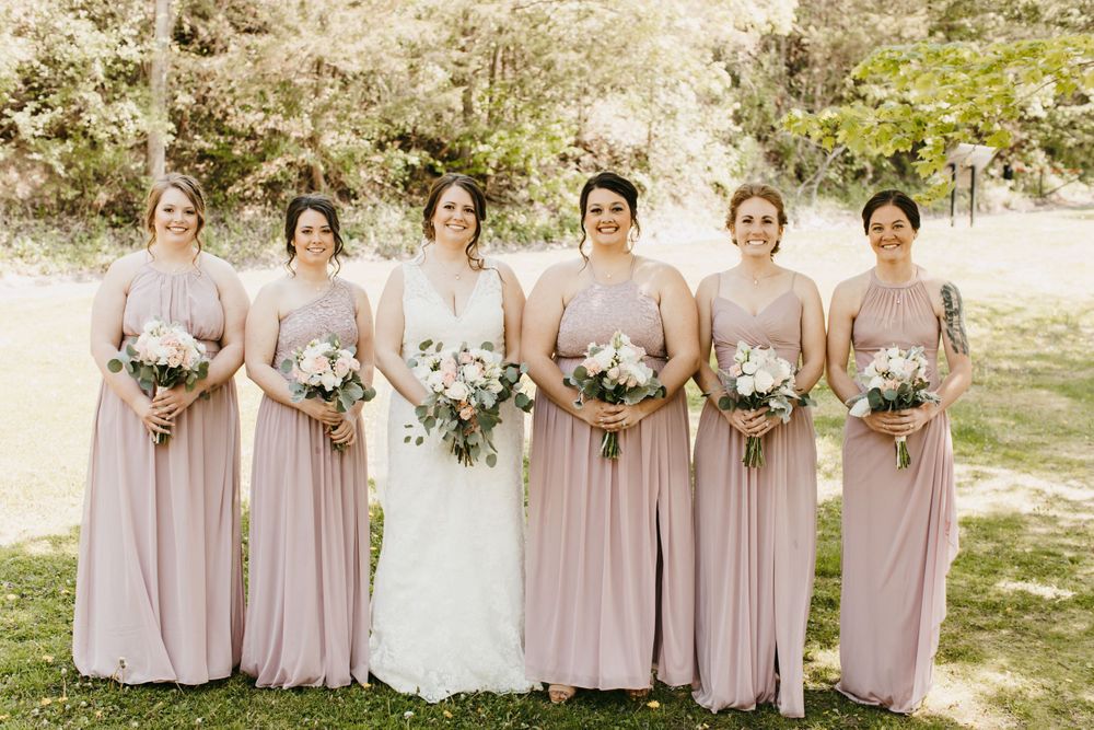 Bride in white lace dress stands with five bridesmaids wearing matching dusty pink gowns and floral bouquets.