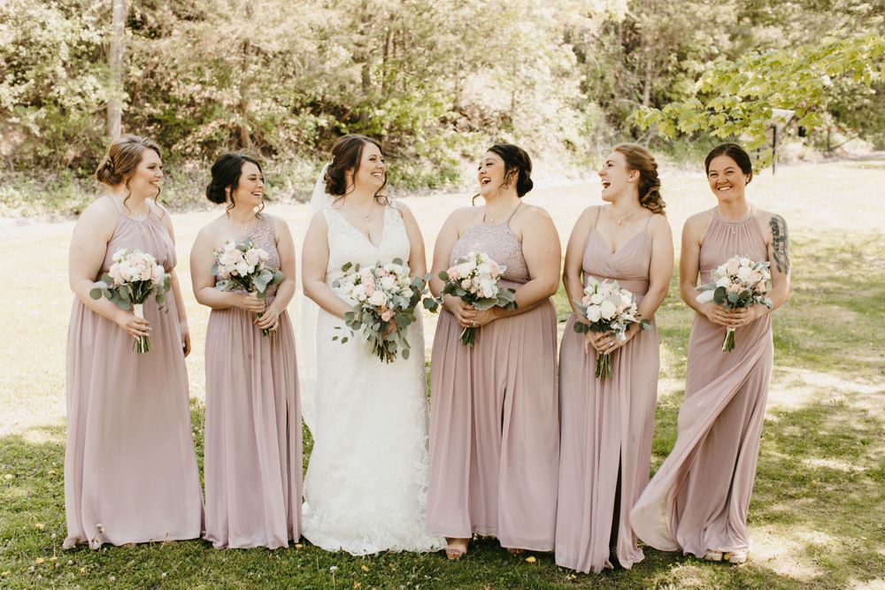 Bride and bridesmaids in dusty rose gowns laugh together while walking outdoors in a lush green setting.