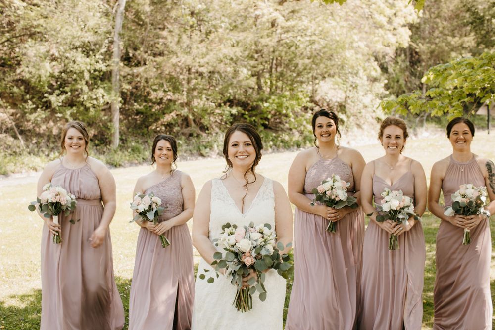 Bride in white gown smiles surrounded by five bridesmaids in mauve dresses holding bouquets outdoors.