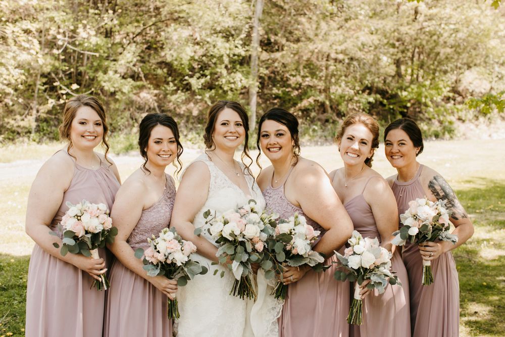 Bride and five bridesmaids in dusty pink dresses smile and hold floral bouquets in an outdoor garden.