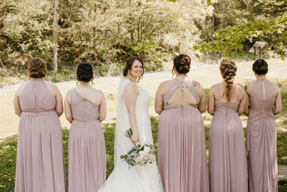 Bride faces camera smiling as five bridesmaids in dusty rose gowns show their backs in an outdoor setting.