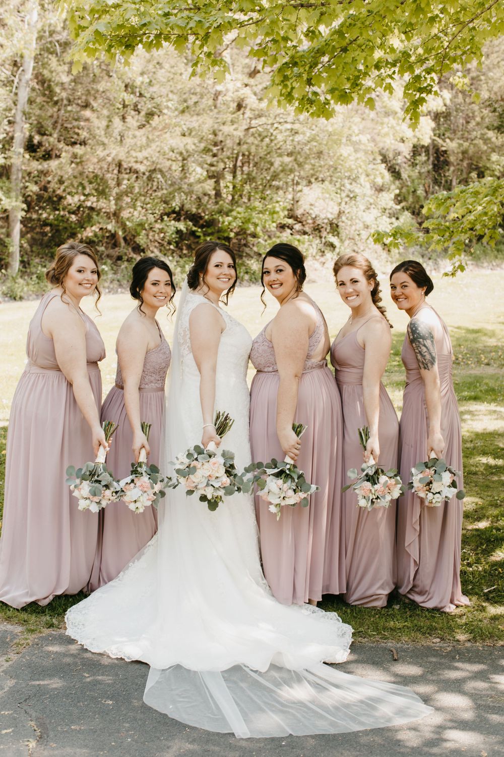 Bride in white gown with long veil poses with five bridesmaids in mauve dresses holding floral bouquets.