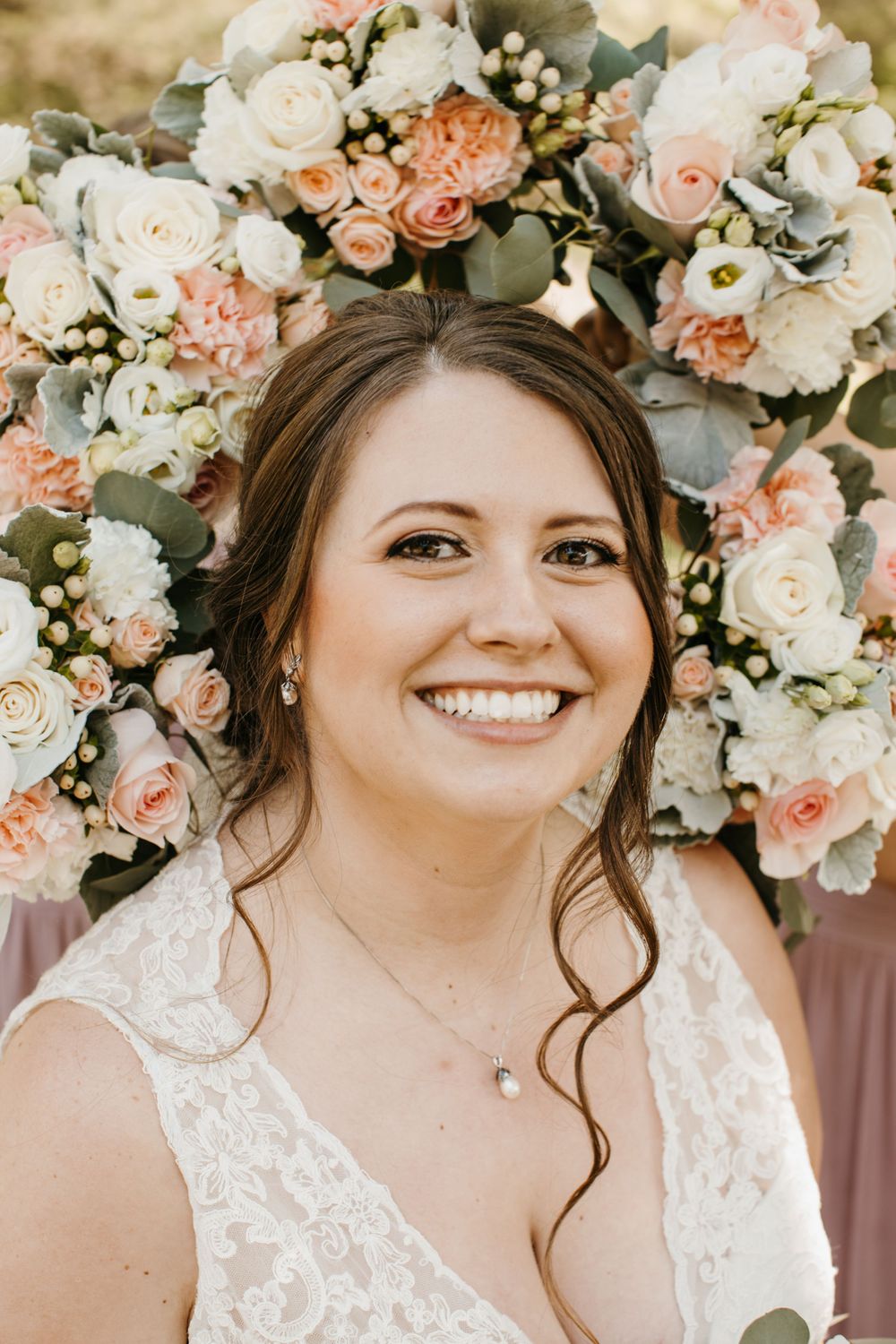 Close-up portrait of a smiling bride in lace gown surrounded by peach and white floral arrangements.