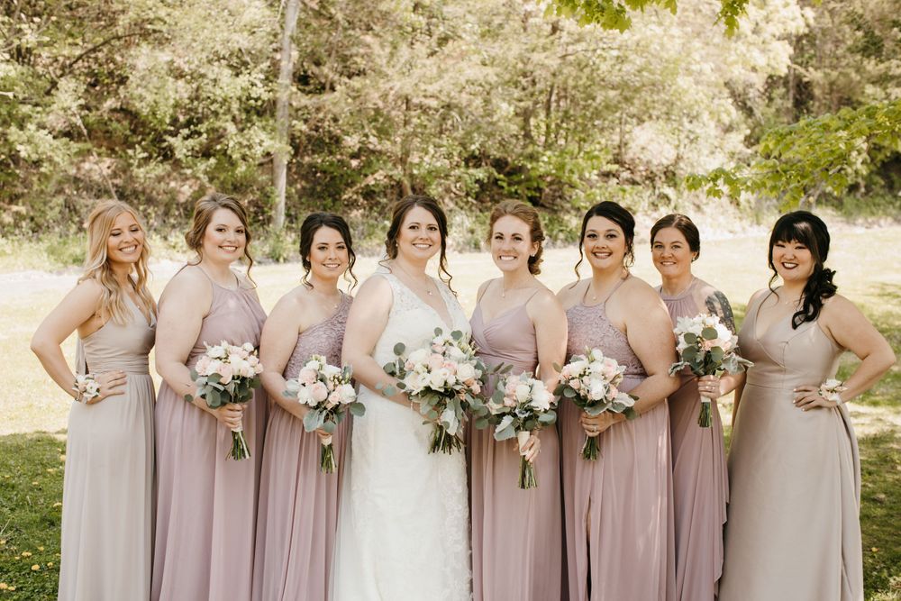 Bride in white dress stands with seven bridesmaids in dusty rose gowns holding bouquets outdoors.