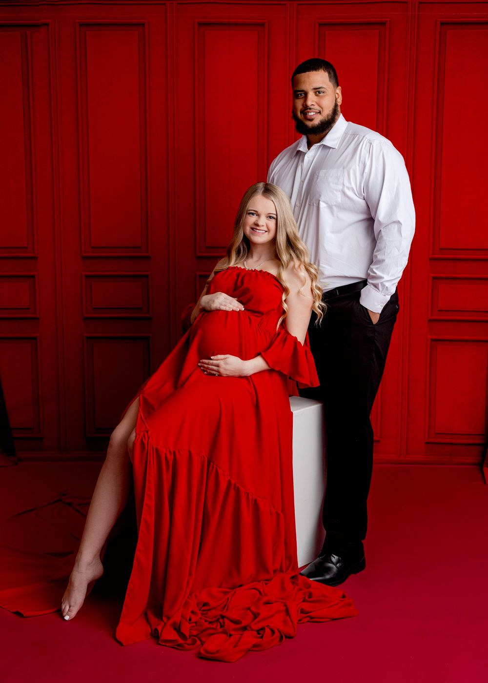Maternity photo of couple in formal attire against bold red paneled wall.