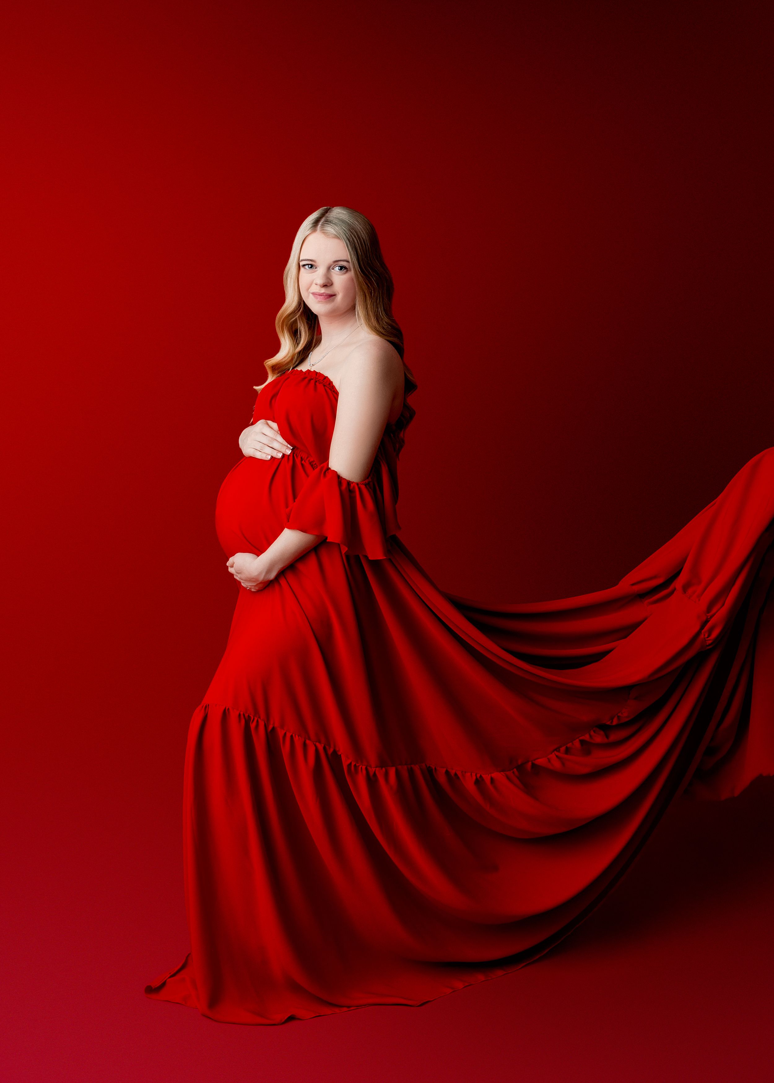 Women with long blonde hair in a red flowing gown holding her pregnant belly at a photography session in Worthington Ohi